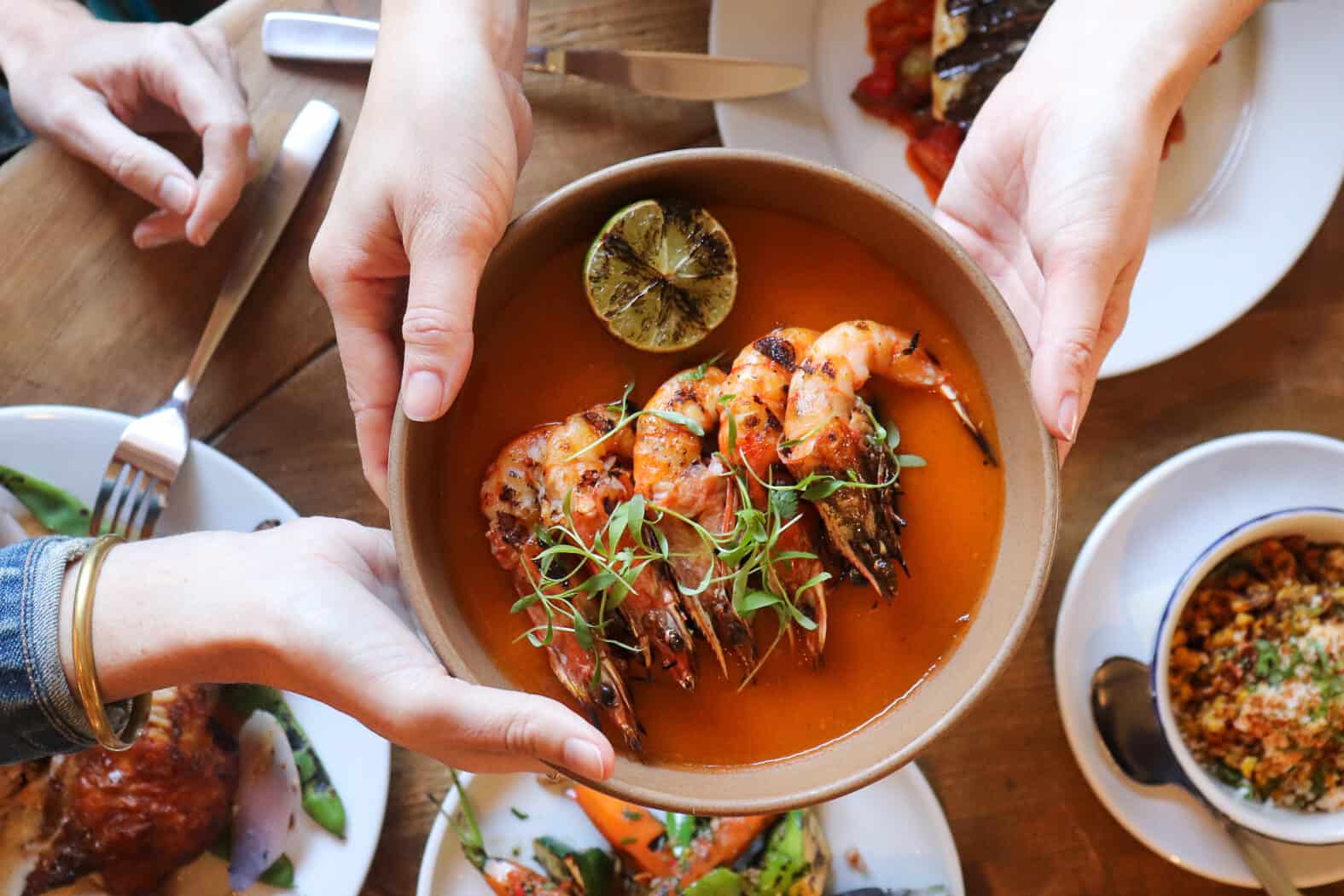 Hands passing Camaron a la Parilla over a spread of food