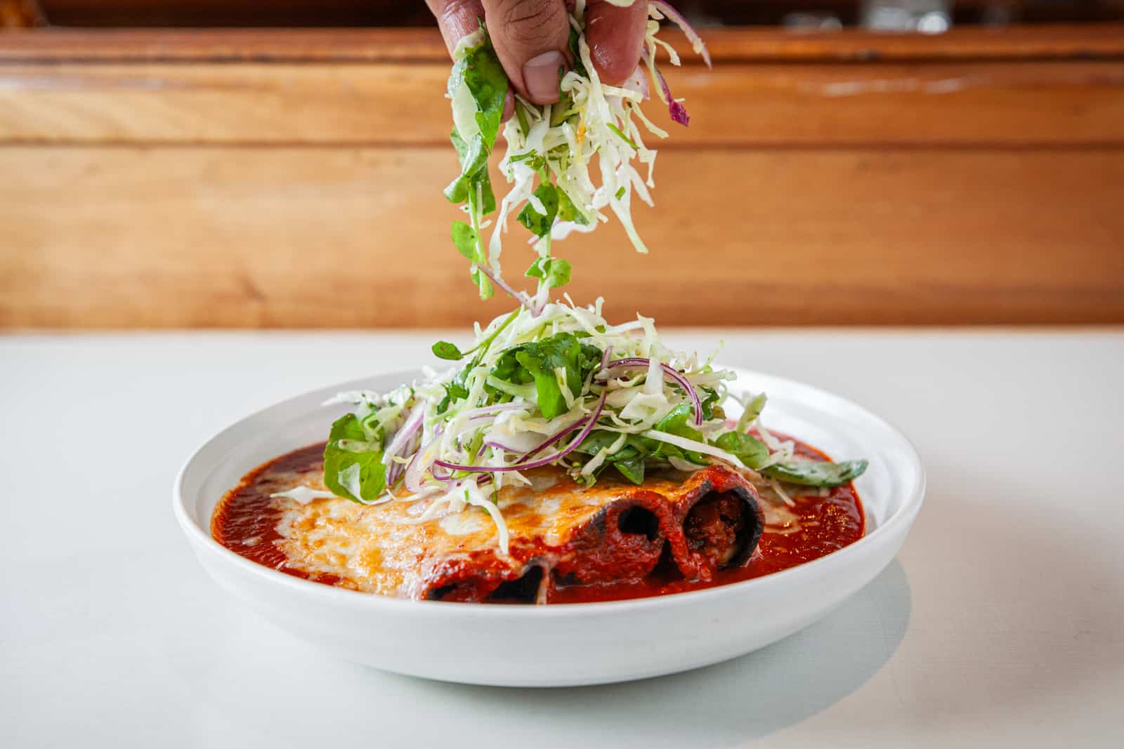 hands garnishing enchiladas in a red sauce with cabbage and micro greens
